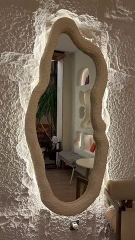 The video opens on a close-up of a large, irregularly shaped mirror with a textured, beige frame. The mirror is mounted on a textured white wall with embedded lighting that creates a halo effect around the mirror. The camera then pans slowly to the right, revealing more of the textured wall, which features a series of organically shaped niches or shelves. These niches are also backlit, creating a soft glow.

In the foreground, to the right of the mirror, is a small, round, light-colored table with wooden legs. Next to the table is a light-colored armchair with wooden arms and legs. Further to the right, a tall decorative dried grass arrangement is visible.

As the camera continues to pan, more of the niche-filled wall is shown. Some of the niches contain decorative items, such as books, small plants, and vases with dried reeds or flowers. In the background, a narrow bench with a gray cushioned top and gold legs is visible, along with a glimpse of a potted plant and some furniture beyond. The floor is made of light-colored wooden planks. The overall impression is of a stylish, modern interior with a focus on organic shapes and soft lighting.
