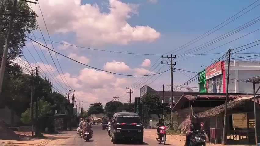 The video depicts a street scene in an unspecified location. The weather is sunny with scattered clouds. A black van filled with people drives down the road, followed by several scooters and motorcycles. The street is lined with buildings and utility poles with numerous wires. One building has a sign that reads "PT. CORIN MULIA GEMILANG." A "Pegadaian" sign is also visible. The overall scene appears to be a typical day on a busy street.