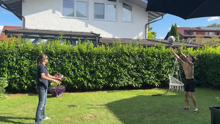 Two people are playing volleyball in a backyard. A woman with sunglasses and a dark t-shirt is on the left, and a shirtless man with dark shorts is on the right. The woman is in the process of hitting the volleyball, which is in the air above her. The man is in a defensive stance, ready to receive the ball. The backyard has a lush green lawn and a tall, dense green hedge that separates it from the neighboring properties. A white house with several windows is visible in the background, along with a portion of a black umbrella. The sky is blue with some clouds, indicating it is a sunny day.