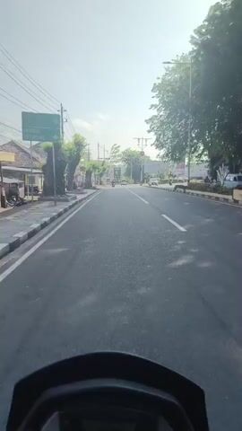The video captures a point-of-view perspective from a vehicle, likely a motorcycle, traveling down an asphalt road. The road is lined with sidewalks and trees on both sides. Buildings and shops are visible along the left side of the road, some with signs. Utility poles and wires are present overhead. Traffic signals are visible at an intersection ahead. Several other vehicles, including cars and motorcycles, are also on the road. The sky is bright, suggesting daytime, and the sun casts shadows. The video appears to be a short recording of a journey through a town or city.