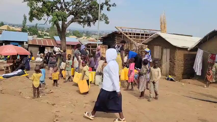 The video shows a group of people, predominantly children and women, gathered in an outdoor area that appears to be a village or settlement. The ground is dirt and uneven. There are several simple structures made of wood, mud, and corrugated metal, some with thatched roofs. Some buildings appear unfinished or damaged. Several large yellow containers, likely for water, are present, and some people are holding them. A few individuals are wearing traditional African clothing, while others are in more modern attire. Some people are walking, while others are standing and observing. The overall atmosphere seems to be one of community gathering, possibly for the distribution of essential resources. The background shows a hilly landscape with more buildings in the distance. The lighting suggests daytime.