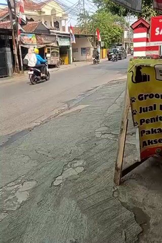 A wide shot shows a street scene. In the foreground, a yellow A-frame sign stands at an angle, partially obscuring the view of the sidewalk. The sign has paw prints and text that reads "GROOM JUAL HA PENITIPAN PAKAN AKSESORIS". In the middle ground, a two-lane asphalt road is visible, with a white line dividing the lanes. A motorcycle is traveling away from the camera in the left lane, with two people on it, one wearing a yellow hijab. Several small buildings line the street on the left, including what appears to be a food stall. On the right side of the street, across from the buildings, there is a red and white striped pole and a red sign with white text. A few trees and power lines are visible in the background. The weather appears to be sunny.