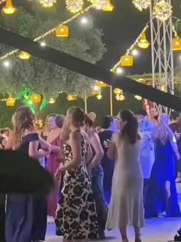 The video shows a night-time outdoor event with many people dancing. Numerous string lights with yellow lanterns and spherical light fixtures are visible overhead, illuminating the scene. A large black boom arm, likely for a camera or microphone, is prominently positioned in the foreground, obscuring parts of the dancers. Several individuals are captured in motion, some in formal attire such as dresses. The overall atmosphere suggests a celebration or party.