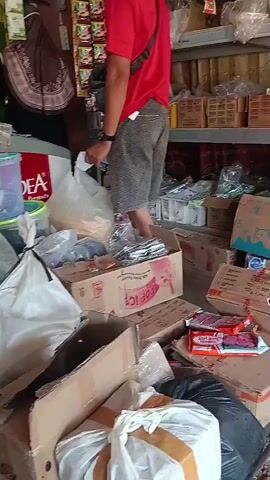 The video pans across a cluttered store. In the foreground, a person in a red shirt and gray shorts stands facing away from the camera, looking at a stack of boxes. To the left, a person in a black hijab sits behind a counter, partially obscured. The store is filled with shelves and stacks of goods, including boxes, bags of products, and containers. Some shelves are stocked with packaged items, and others have loose items. A white refrigerator is visible in the background. The overall impression is of a busy, somewhat disorganized retail environment.