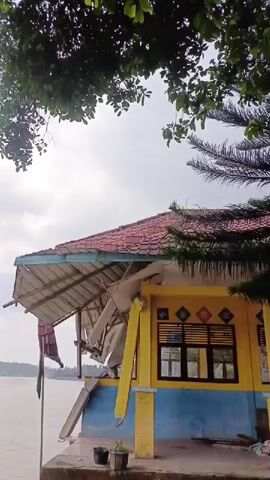 The video captures a riverside scene, panning slowly from right to left to reveal more of the waterway. In the foreground, a colorful building with yellow walls, blue trim, and a red-tiled roof stands directly beside the river. The building appears to be in disrepair, with a significant portion of its front porch or extended roof structure having collapsed or severely damaged. Wooden beams and roofing material are visibly broken and hanging down. Below the damaged section, two small plant pots sit on paved ground, which transitions into a rocky, unkempt bank leading to the water.

The river itself is wide and appears murky brown, stretching into the distance. The opposite bank is lined with dense green trees and vegetation. The sky is overcast with a blanket of white and grey clouds, indicating daytime. No people are visible in the shot. The scene suggests a location where buildings are susceptible to natural elements, possibly river erosion or past severe weather.