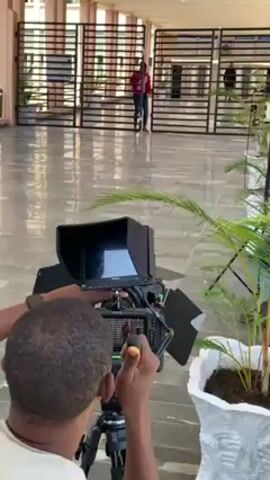 A man is filming a woman with a video camera on a tripod. The man is wearing a white shirt and has short dark hair. He is looking at the camera monitor as he adjusts the camera. The woman is standing in front of a building with a gated entryway. She is wearing a pink jacket and blue jeans. There is a potted plant to the right of the camera. The camera has a monitor and a sun shield attached. The camera is pointed at the woman. The ground is a shiny light gray tiled surface.