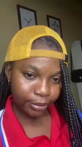 A young Black woman with braided hair, wearing a yellow mesh baseball cap and a red and blue shirt, looks directly at the camera. The background includes framed pictures on a beige wall. The woman is indoors.