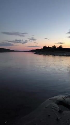 A wide, calm river flows at dusk. The sky is a gradient of deep blue at the top, fading to soft pink and orange near the horizon. Wispy clouds are scattered across the sky. Silhouetted trees and a distant landscape line the far bank of the river. The foreground shows a sandy bank with a small pool of water. The lighting suggests it is either sunrise or sunset. No people or specific actions are visible.