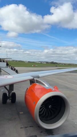 The video begins with a close-up of an orange jet engine on an airplane, with passengers visible on a boarding ramp in the background under a cloudy sky. The scene transitions to a view from inside the aircraft as someone walks down the aisle. The camera then focuses on a man sitting in a seat, wearing glasses, a black t-shirt, and jeans, looking around while holding his phone. The environment appears to be inside a fully occupied commercial airplane.