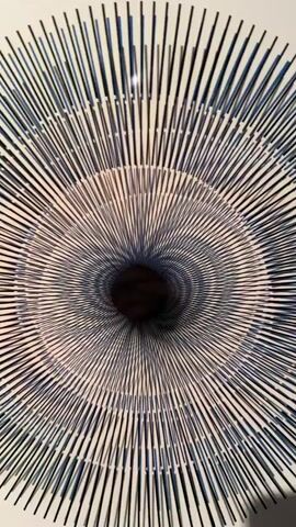 This is a close-up shot of a swirling, hypnotic pattern. The pattern is made up of thin, vertical lines radiating outwards from a dark, central void, creating a spiral effect. The lines appear to be blue and white, and they create a sense of depth and movement as they recede into the darkness. The lighting is soft, casting a gentle glow on the pattern.