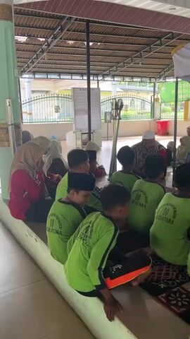A group of children wearing green shirts with the text "MADRASAH IBTIDAIYAH NEGERI 10 ACEH UTARA" are sitting on the floor facing an older man and a few other adults. The children are wearing black skullcaps. The older man is wearing a white skullcap and a patterned shirt. They are seated under a covered area with a corrugated metal roof. In the background, there is a tomb or monument with a stone structure. Some of the adults are wearing headscarves. The environment appears to be outdoors, possibly a courtyard or a religious site. The children appear to be listening to the older man.
