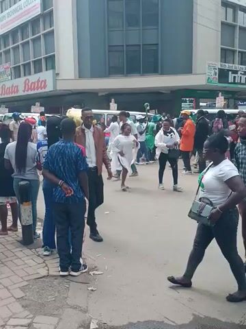 The video shows a group of people walking along a street. The buildings in the background have signs for Bata and a Technical College. Some people are dressed in white, while others wear a variety of colors and patterns. The camera appears to be moving along with the group. The street appears to be paved.