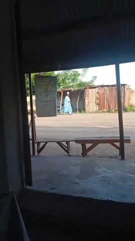 The video begins with a view of an open doorway. Outside, a person in a blue garment is walking away from the camera. Behind them, there are some corrugated metal structures and vegetation. The camera then pans inside to show a desk with electronic equipment and a person wearing a cap. The video ends with another view of the outside scene through the doorway.