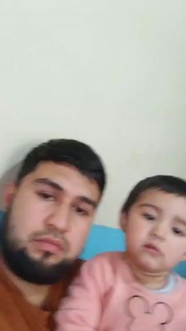 A video shows a selfie with a father and a young child. The father is on the left side of the frame, with dark hair and a beard, wearing a brown shirt. The child, on the right, has short, dark hair and is wearing a pink sweater with a black outline of Mickey Mouse. The background is a plain off-white wall with a hint of blue behind them. The camera remains still, capturing their upper bodies. The child is looking at the camera.