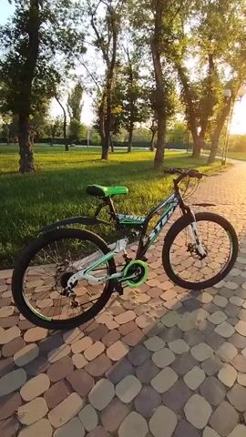 A bicycle rests on a paved walkway in a park. The sun is setting, casting long shadows and illuminating the scene with a warm glow. Trees line the walkway, and a grassy area is visible to the left. The bicycle is black and white with green accents. The camera pans slowly to the right, capturing the tranquil park setting and the setting sun.