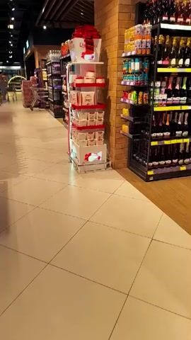 The video shows a stationary point-of-view shot within a well-lit supermarket aisle. The aisle features prominent shelving units displaying a variety of products. To the left, a display stand with red and white packaging, possibly confectionery or gift items, is visible. Beyond this, another aisle with more shelving is suggested. To the right, a tall, black metal shelving unit is filled with numerous bottles of various sizes and colors, primarily appearing to be wine or spirits. The bottles are arranged neatly on multiple shelves. The floor is tiled with large, light-colored square tiles, and the walls have a textured brick finish on the right side of the frame. The overall environment suggests a retail setting, likely a liquor store or a supermarket with a dedicated alcohol section. No people are visible in the frame. The lighting is bright and consistent, illuminating the products and the store environment.