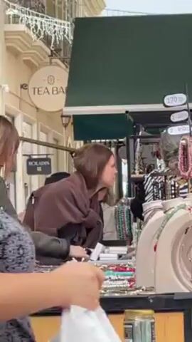 The video shows several women at an outdoor jewelry stand, on what appears to be a European street. A woman in a green jacket and plaid skirt looks at jewelry on the table. Another woman with brown hair wrapped in a scarf stands next to her, also looking at the jewelry. Behind them, a woman in a black jacket looks at the jewelry. Another woman in a zebra print shirt is behind the jewelry stand, presumably working there. The background shows buildings with decorative details, a Tea Bar sign, and string lights overhead. The sounds of a band playing can be heard.