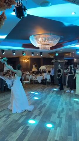 A bride in a white wedding dress stands in a ballroom, with her arm raised to throw a bouquet of flowers. She is facing away from the camera, and a group of women are gathered to her right, presumably waiting to catch the bouquet. The room is lit with colorful lights, and a large chandelier hangs from the ceiling. Tables and chairs are set up for guests. The floor is made of wood.