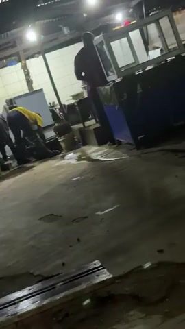 The video shows an indoor scene, possibly a market or food stall, with a cracked concrete floor. Two individuals are visible, one wearing a yellow shirt and the other in dark clothing. They appear to be interacting or working near a food display laden with fruits like watermelons, grapes, and apples. A third person is standing near a cooler in the background. The lighting is dim, provided by overhead fluorescent lights. The camera's perspective is low and slightly angled, and the footage is shaky.