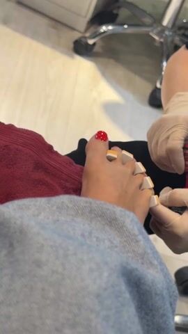 A pair of feet is seen in a pedicure chair, with toe separators in place. The big toe of one foot is painted red and glittery. A person in gloves is painting the rest of the toenails with red nail polish. The person is wearing a pair of jeans, and a red towel is visible. The room appears to be a nail salon.