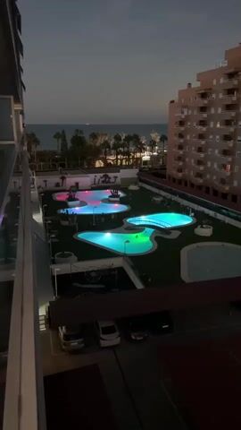 A high-angle, medium shot captures a resort-style courtyard at dusk. Several irregularly shaped swimming pools are illuminated with vibrant colored lights in shades of blue, pink, and green. Palm trees and other vegetation dot the landscape around the pools. In the background, a dark ocean meets a hazy sky, with lights from buildings and possibly a distant shore visible. To the right of the courtyard, a multi-story building with many lit windows looms over the scene. In the foreground, a portion of a balcony railing from the vantage point is visible on the left. Below the courtyard, a covered parking area with several cars is discernible. The overall ambiance is tranquil and resort-like, with the colorful lighting creating a visually striking effect against the dimming natural light.