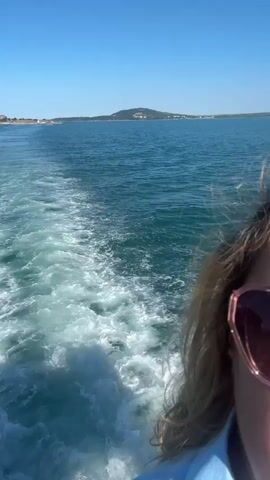 This is a vertical video captured from a boat moving through the water. The foreground is dominated by the wake of the boat, a frothy trail of white water churned up by the propeller. To the right, a person's head and shoulders are visible, partially out of focus. They are wearing large, rose-tinted sunglasses and their blonde hair is catching the sunlight.

The midground shows the expanse of the sea, a vibrant blue under a clear sky. Further in the distance, a coastline is visible with rolling hills covered in greenery and some structures, suggesting a populated area. The sun appears to be high in the sky, indicating it is likely midday. The weather is clear and sunny, ideal for a boat trip. The overall mood is one of leisure and enjoyment, implied by the sunglasses and the presence of a person on a boat ride. The location is identified as Atiya, Bulgaria. There is no visible text in the video.