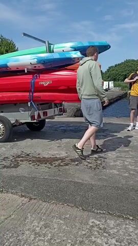 The video shows a group of people standing near a trailer loaded with kayaks. The trailer is parked on a concrete surface, and a body of water is visible in the background, with hills and trees on the opposite shore. The kayaks are stacked on the trailer, with the topmost ones being blue and green, followed by orange, and then red kayaks at the bottom. One person, wearing a green hoodie and grey shorts, is standing with their back to the camera, facing the other people. Two other individuals, one in a yellow shirt and hat, and another in a light-colored shirt and black shorts, are conversing. A fourth person, wearing a green life vest and a hat, is also present. The sky is blue with scattered clouds. The trailer has a single wheel visible on the left side. The overall scene appears to be an outdoor recreational setting, possibly preparing for a kayaking activity.