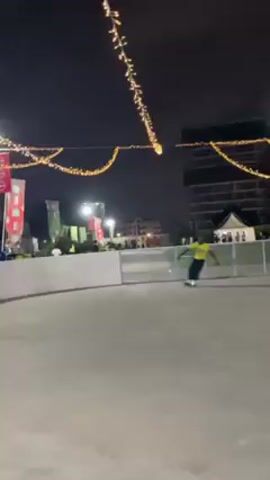 A man in a yellow shirt and dark pants is skating on a paved surface, likely an ice rink, at night. The area is illuminated by strings of fairy lights overhead. He skates towards the camera initially, then turns and skates away, passing near a fence and other people standing by the side of the rink. There are buildings and other decorations visible in the background under the dark sky. The scene appears to be festive and recreational.