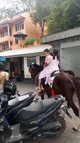 Two young girls ride a brown horse on a street. The girl in front is wearing a pink and white striped dress, and the girl behind her is wearing a pink dress. They are both looking forward. The horse is wearing a red saddle blanket. In the background, there are several shops and buildings, and a few motorcycles are parked on the side of the road. The weather appears to be overcast.