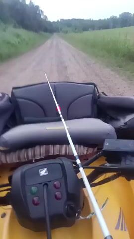 The video shows the point of view from the front of a yellow Honda ATV traveling down a dirt road lined by fields and trees. A fishing rod with a red bobber is mounted on the back seat. The weather appears overcast.