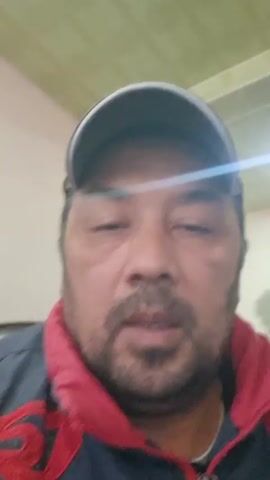A middle-aged man wearing a dark blue jacket with red trim and a baseball cap greets the viewer. He raises his hand and waves at the camera. He appears to be indoors in a room with a light-colored ceiling. The man smiles and rests his chin on his hand.