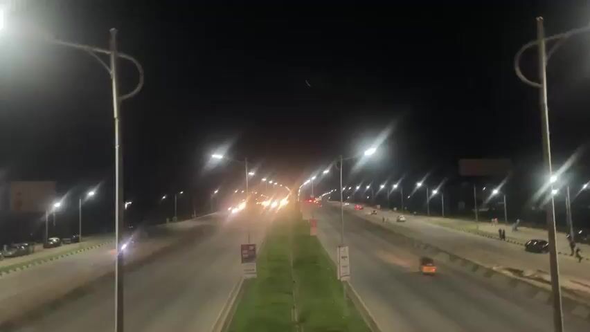 The video is a night shot of a highway. Streetlights illuminate the road and the surrounding grassy median. Cars with their headlights on are driving on the highway. Some cars are parked on the side of the road. The sky is dark.