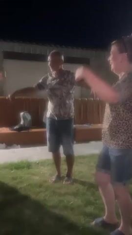 Two adults are dancing outdoors at night. A male with short hair and a patterned t-shirt and denim shorts stands on the left, his arms extended to his sides. A female with blonde hair pulled back and a leopard-print t-shirt and denim shorts is on the right, her arms raised and bent at the elbows. They are on a grassy area with a wooden bench and some objects on it in the background. The scene is dimly lit.