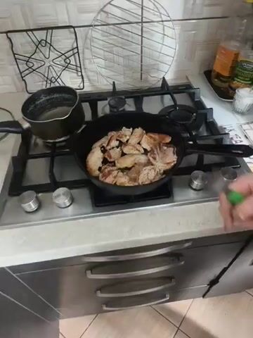 A close-up, slightly shaky, overhead shot shows chicken pieces being fried in a black cast-iron skillet on a gas stove. The chicken pieces are unevenly cut and varied in size. Some pieces are lighter in color, while others are browning, indicating they are in different stages of cooking. The skillet is on a stainless steel stovetop. A white tiled backsplash is visible behind the stove. On the counter to the right, a few items are partially visible, including a glass jar with a yellow lid and a white shaker. A person's hand holding a green-handled utensil, possibly a spatula or tongs, is visible at the bottom right of the frame, implying they are tending to the cooking. The lighting is typical of an indoor kitchen environment.