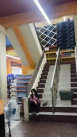 A person sits on the stairs in a store. The person is wearing a pink hoodie and black pants. The person has black hair and is wearing sunglasses. There are shelves with products in the background. There is a fan in the foreground.