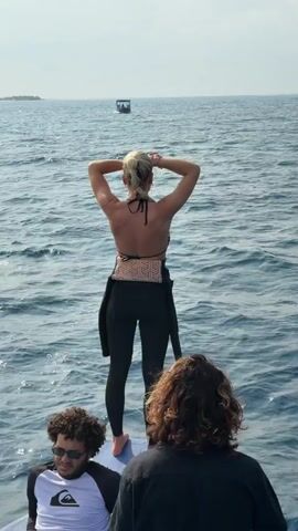 The video shows three people on a boat in the ocean. A woman with blonde hair stands at the front of the boat, wearing a zebra-striped bikini top and black pants. She adjusts her hair and looks around. Behind her stands a man with long, curly hair, also adjusting his hair. To the left, another man sits wearing sunglasses and a white long-sleeved shirt with black sleeves. The ocean is a clear, light blue, and a small building can be seen on the horizon. The sky is overcast. The boat is moving, creating ripples in the water.