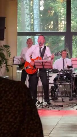 A band plays music on a stage. Three men are visible. The man in the center is singing and playing an orange electric guitar. He is wearing a white button-down shirt, a black tie, and black pants. To his left, another man stands behind a music stand, also wearing a white shirt and black pants. To the right of the center man, a third man is seated behind a drum set, wearing a white shirt and a black tie. He is looking down, possibly playing the drums. The stage is lit with red and white lights. Behind the band, a large window overlooks a wooded area with trees. A large speaker is visible to the left of the stage. The foreground is dark and out of focus, with a hint of textured fabric.
