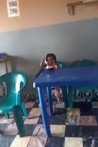 A young girl with dark braided hair is seated at a blue rectangular table, eating from a bowl with a spoon. She is wearing a light-colored top and pink pants. The table is supported by four blue legs. Two green plastic chairs are visible, one on the left and another behind the girl. The floor is tiled with a black and white checkered pattern. The wall behind the girl is painted in two horizontal bands, gray on top and a lighter color below. A light-colored sign with text is affixed to the upper wall on the left.