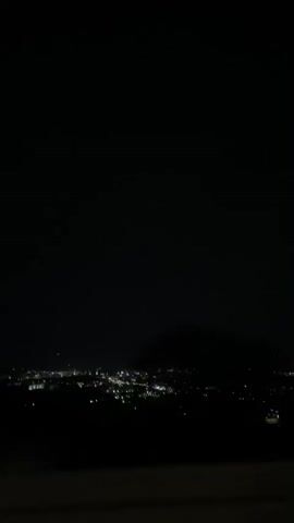 The video shows a night cityscape from a high vantage point, with city lights twinkling below against an otherwise dark sky. Some cloud cover is visible. The view seems to be from a stationary position.