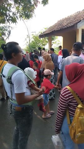 This media file shows a group of people, including adults and children, gathered outdoors. The scene appears to be in a village or community area in Posong, Indonesia, as suggested by the traditional Indonesian architecture of the building visible in the background, and the presence of people in modest attire, with many women wearing hijabs.

The subjects are diverse, with individuals of various ages and genders. Some people are dressed in casual clothing, while others wear more athletic attire, suggesting a possible community event or celebration. One prominent group in the foreground includes a man carrying a child in a sling, a young boy in a red shirt and shorts with Indonesian text, and a young girl wearing a yellow hat. Behind them, a woman in a red hijab and striped shirt is visible.

The activity appears to be a gathering or a procession. People are standing and walking, with some engaging in conversation. The atmosphere seems cheerful and communal, with a sense of shared activity. The time of day appears to be daytime, likely morning or afternoon, given the natural light and the shadows. The weather seems pleasant, as people are outdoors without heavy jackets.

Notable details include the building in the background, which has a tiled roof and a light-colored exterior, suggesting it could be a community hall or a small office. There is also greenery and trees surrounding the area, indicating an outdoor setting. A long concrete structure, possibly a bench or a low wall, runs along the path where people are gathered.

Visible text on the clothing of the young boy reads "DIRGAHAYU REPUBLIK INDONESIA," which translates to "Long Live the Republic of Indonesia." This text strongly suggests that the event is related to Indonesian Independence Day or another patriotic celebration. The presence of multiple families and children indicates a community-focused event.