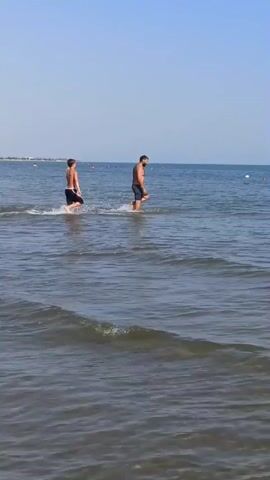 Two males walk through shallow water near a beach. The male on the left is younger and thinner. He is wearing dark swim trunks. The male on the right is larger and has a beard. He is also wearing dark swim trunks. The water is clear and blue. The sky is clear and blue.