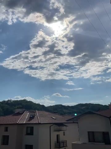 The video captures a panoramic view of a town nestled in a valley, surrounded by lush green hills. The sky is a mix of blue and white clouds, with the sun intermittently peeking through, casting rays of light. The architecture of the town appears to be a blend of older, traditional buildings with red-tiled roofs and some more modern structures, including apartment buildings. The foreground features the rooftops of several houses, some with visible chimneys. The overall impression is one of a peaceful, somewhat rural town under a dynamic sky. There are no people or animals visible in the video. The camera appears to be stationary, observing the scene unfold.