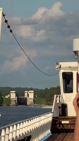 The video shows a view from the deck of a ship approaching a canal lock system. The lock gates are visible in the distance, appearing to be open or partially open. The ship has a white railing with blue trim. A white pole with a taut wire is visible on the left side of the frame. The sky is partly cloudy. Trees and buildings are visible on the shore behind the lock. The water in the canal appears calm.