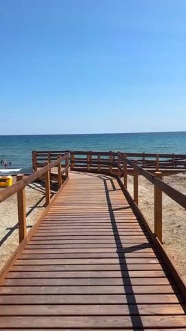 A first-person perspective video shows a wooden boardwalk curving towards the sea. The boardwalk is made of light brown planks and has wooden railings on both sides. The sea is visible in the background, with clear blue water and gentle waves. The sky is a bright, cloudless blue. There is a sandy area next to the boardwalk. A few people are visible in the water near the shore. The sun is casting shadows of the boardwalk onto the sand.