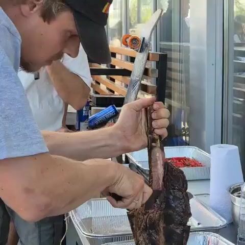The video shows a person wearing a grey t-shirt and a black baseball cap with a logo, carving a large piece of meat on a metal table. The meat, which appears to be a prime rib roast or a similar cut with a bone, is held vertically by the bone while the person uses a knife to slice off portions. The meat has a dark, cooked exterior and a pink, raw interior. The person is focused on the carving process. In the background, there are several metal pans, one of which contains what looks like red peppers. There is also a roll of paper towels, a plastic bottle of water, and other containers on the table. The environment appears to be outdoors or in a semi-outdoor setting, with a wooden structure and what looks like glass windows or doors in the background. Another person, wearing a white t-shirt, is partially visible to the left. The lighting suggests daytime.