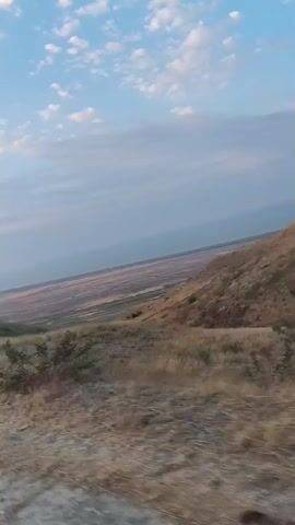 This is a first-person perspective video of a vehicle driving on a dirt road. The road is rocky and uneven. To the left of the road is a dry, grassy hillside. In the distance, there is a wide, flat valley with some sparse vegetation. The sky is light blue with some scattered white clouds. The sun appears to be low in the sky, suggesting it may be sunrise or sunset. The overall scene is one of a dry, rural, and somewhat barren landscape.