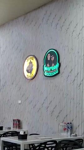 The video shows the interior of what appears to be a cafe or restaurant. The walls are paneled with vertical white slats, and a patterned wallpaper with diagonal lines is visible on the left side of the wall. Two illuminated signs are mounted on the wall. The left sign is round and features a depiction of a drink. The right sign is green and oval-shaped with neon lettering that says "HERKOP" and some smaller text underneath.

In the foreground, several white tables with black chairs are set up. Some tables have black napkin dispensers and bottles of water. In the background, a red counter is visible, behind which a stack of canned drinks is arranged in the shape of a pyramid. To the right of the pyramid, more canned drinks are stacked in rectangular formations. A glass display case sits on top of the counter. A man is standing behind the counter, appearing to be interacting with the canned drinks. The ceiling is white and appears to be a suspended ceiling with recessed lighting fixtures.