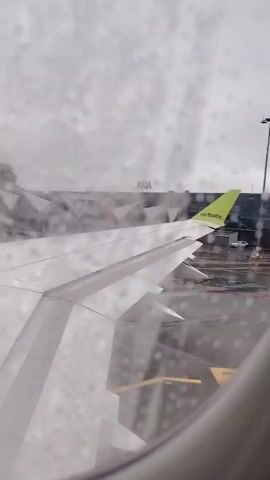 A stationary, low-angle, medium shot from inside an airplane shows the plane's wing parked on the tarmac in Riga, Latvia, with the city's airport in the background. The plane's wing, visible from the window's perspective, is white with a chartreuse green tip. The weather is rainy, with visible raindrops on the window. The airport buildings in the background are light gray, and a control tower is visible behind the plane's wing. The ground is wet and reflects the plane's lights. The sky is overcast and gray.