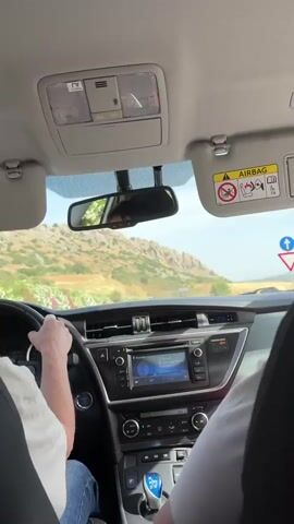 The video is shot from the perspective of a passenger in the front seat of a car. The driver's hands are visible on the steering wheel. The dashboard of the car is in focus, with the infotainment screen displaying a music player. In the foreground, a person's feet are propped up on the dashboard, their toes wiggling. Outside the car, a winding road can be seen, with hills covered in vegetation and scattered rocks. The sky is bright, suggesting daytime. The interior of the car appears to be a typical modern vehicle with sun visors and a rearview mirror. The overall impression is one of a leisurely drive through a scenic landscape.