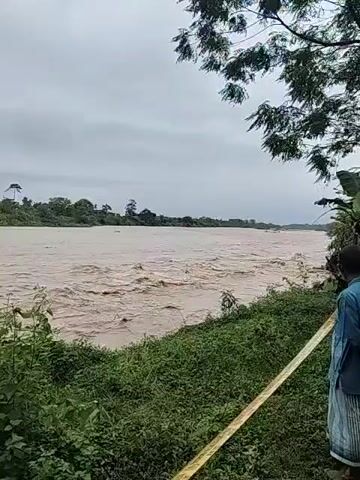 The video shows a muddy river flowing rapidly. A group of people are standing on the riverbank, watching. The riverbank is grassy with trees. A yellow barrier tape is visible in the foreground. The sky is overcast. Some people have umbrellas. There is a building visible further away on the riverbank.